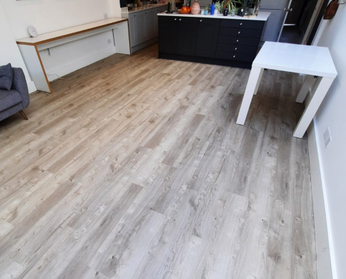 Light sun bleached oak LVT flooring in Reigate kitchen diner viewed from dining area towards grey kitchen cabinets and navy blue island