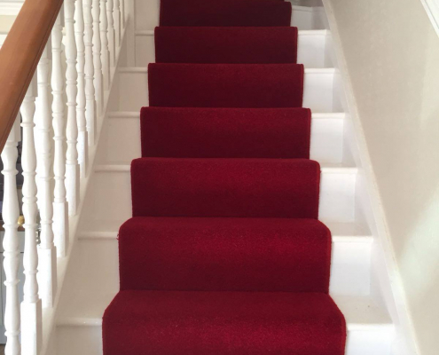 Red wool carpet stair runner on white painted staircase with oak handrail and bannister viewed from bottom looking up