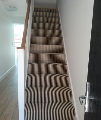 Black and cream striped wool loop carpet staircase viewed from ground floor in bright hallway with white walls and oak handrail