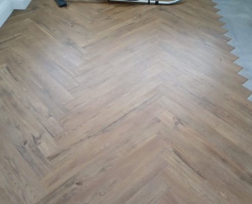 Mid installation of Amtico warm oak LVT flooring in a herringbone pattern with three sections laid over a prepared concrete subfloor in a bright Redhill living room