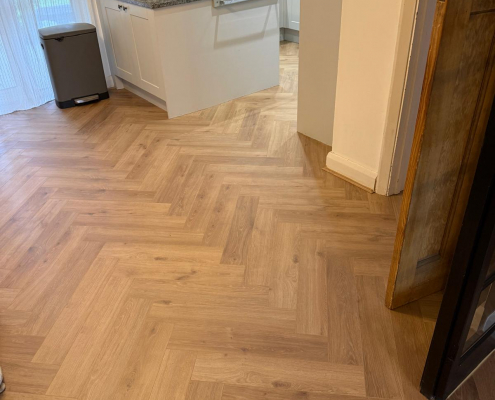 Open Plan Dining Area with Herringbone Laminate Flooring Dining area with warm oak effect herringbone laminate flooring viewed from the living room, featuring white walls and black framed glazed sliding doors.