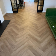 Detailed view of Furlong Manor warm oak effect herringbone laminate flooring installed in bright living room with white walls and natural light.