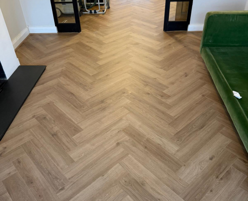 Warm Oak Effect Herringbone Laminate in Living Room Detailed view of Furlong Manor warm oak effect herringbone laminate flooring installed in bright living room with white walls and natural light.